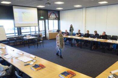 Een docent geeft een training terwijl deelnemers aandachtig luisteren aan de tafels eromheen